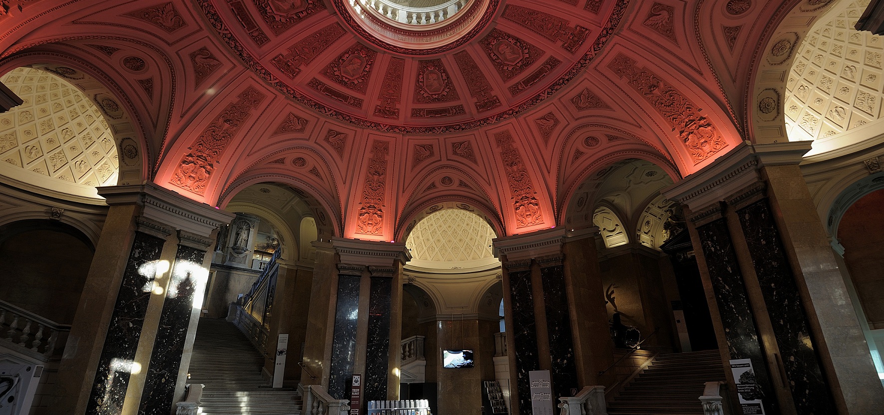 Große Kuppelhalle mit roter Beleuchtung, Marmorsäulen und breiter Treppe in einem historischen Gebäude.