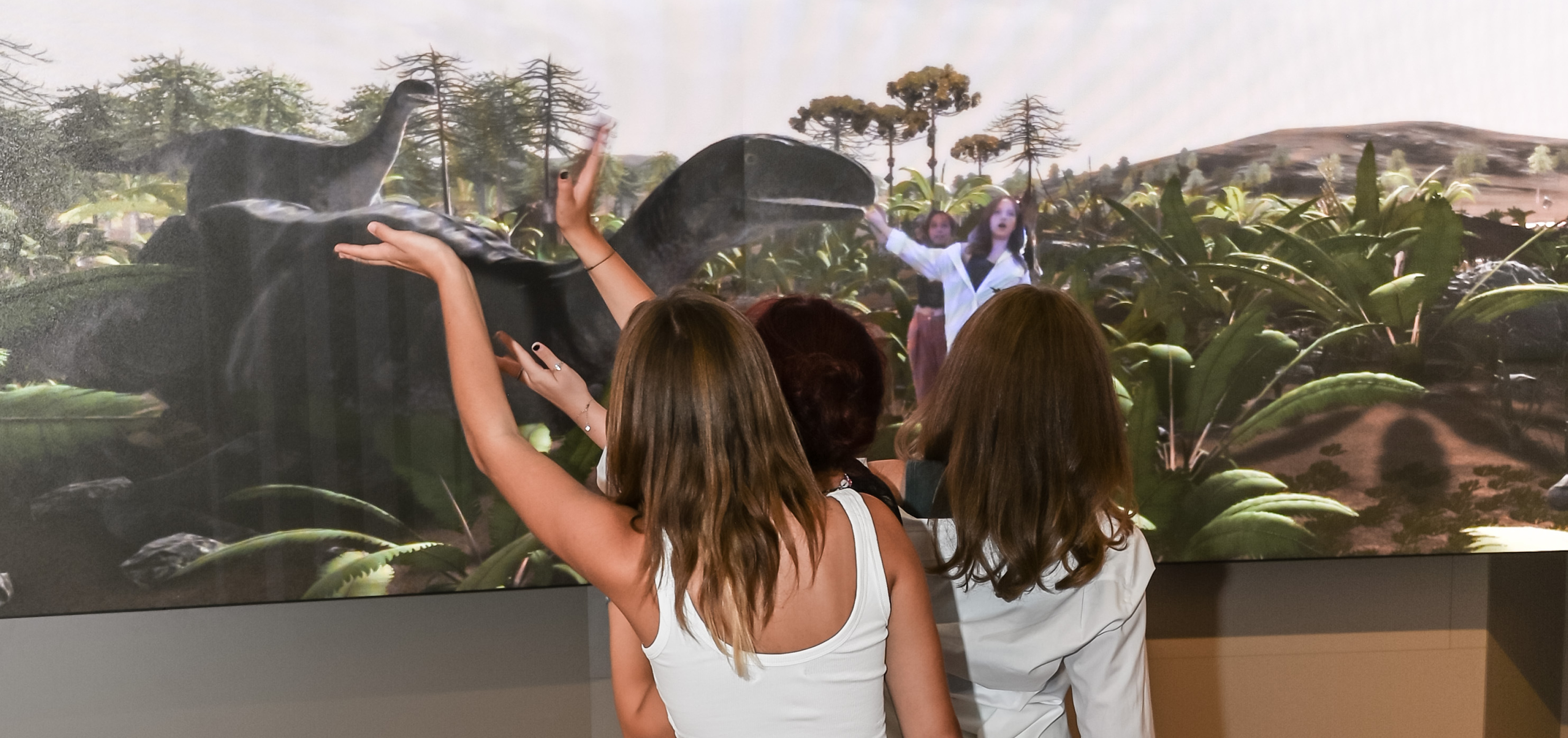 Dinoshow_©NHM Wien_Christina Rittmannsperger Drei Frauen von hinten, die vor einer Dinosaurier-Animation auf einer LED-Wand stehen und mit ausgestreckten Armen Bilder von eines Dinosauriern betrachten und dabei via Kamera in die Landschaft eingespielt werden