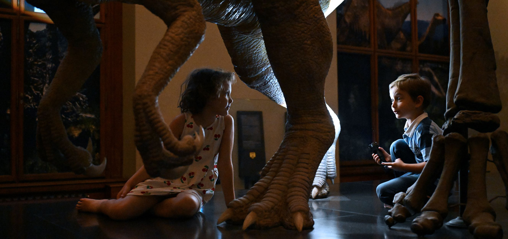 Nacht_Kinder_©NHM Wien_Kracher Nacht Kinder Nacht_Kinder_©NHM Wien_Kracher A Night at the Museum for Children_©NHM Wien_Kracher Zwei Kinder sitzen auf dem Boden unter einem Dinosaurierskelett im NHM Wien