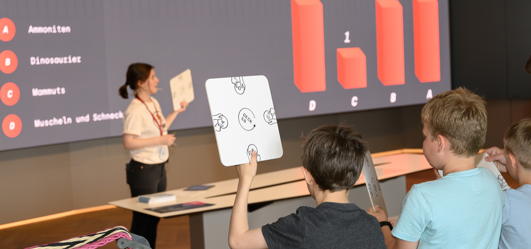 Quizshow_©NHM Wien_Christina Rittmannsperger Kinder halten quadratische Quizkarten hoch, Moderatorin steht vor großer Leinwand mit Frage zu Lieblings-Fossilien und Balkendiagramm der Antworten