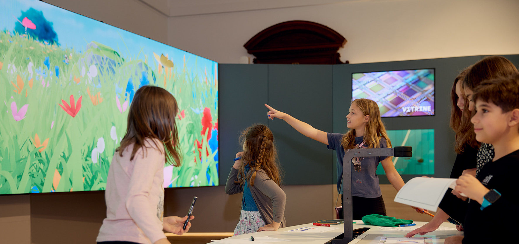 Ferienspiel_D50_©Haeusler Kinder betrachten und interagieren mit einer großen, bunten digitalen Wandprojektion mit Blumen und Schmetterlingen in einem Ausstellungsraum