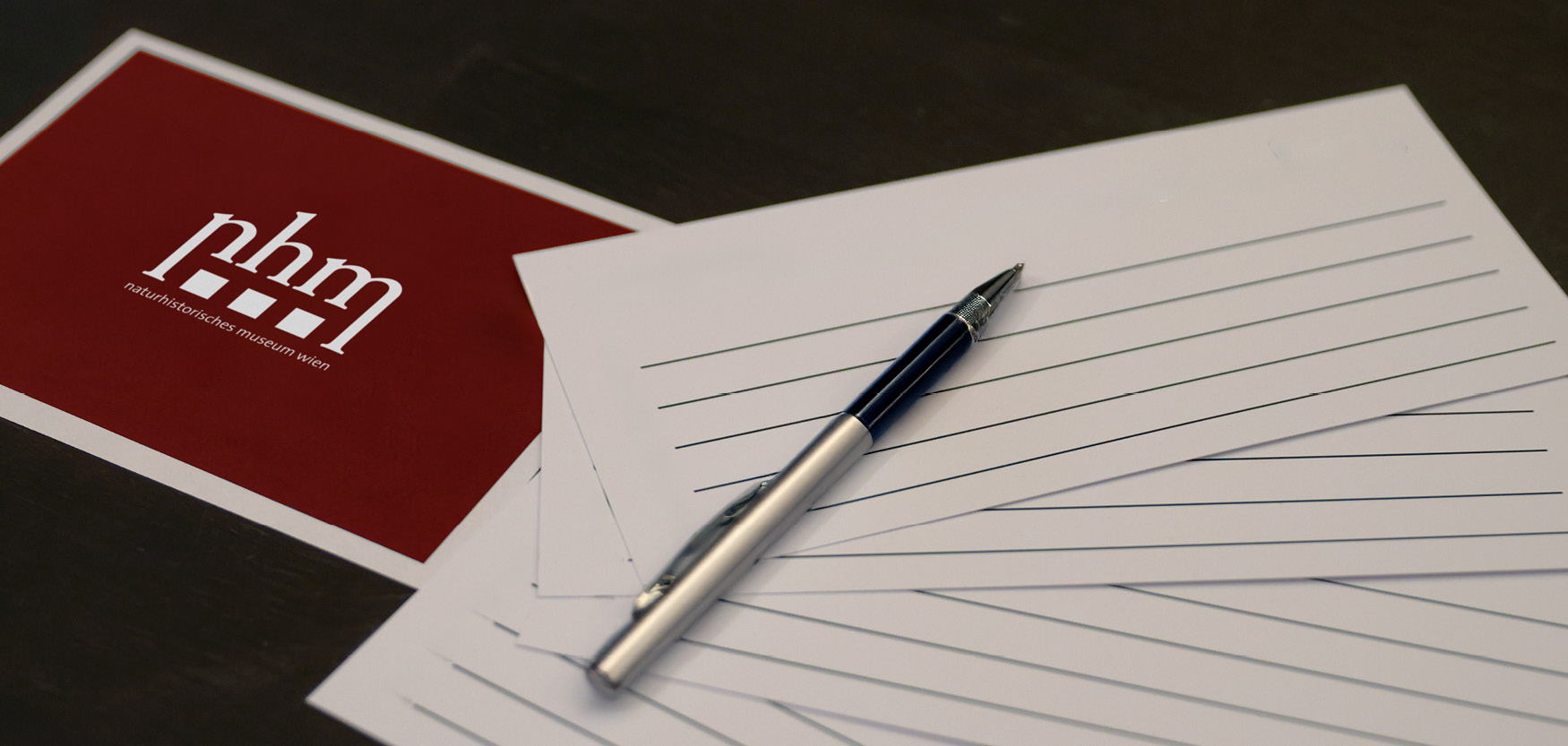 Kugelschreiber liegt auf mehreren linierten weißen Blättern neben einer roten Karte mit weißem Logo und Text
