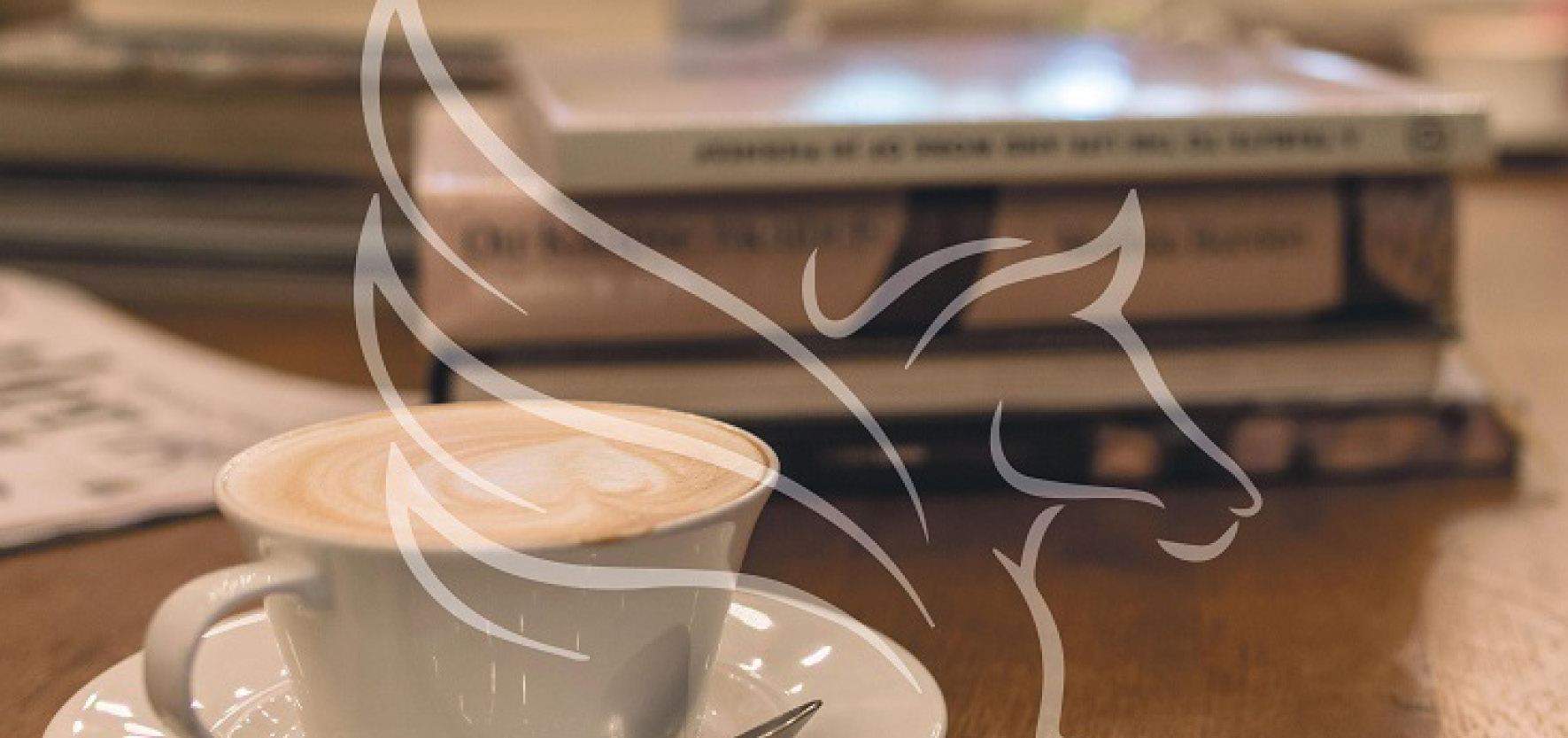 PoetischesFrühstück_©NHM Wien Weiße Tasse mit Cappuccino und Milchschaum auf Untertasse mit Löffel, im Hintergrund mehrere Bücher auf Holztisch