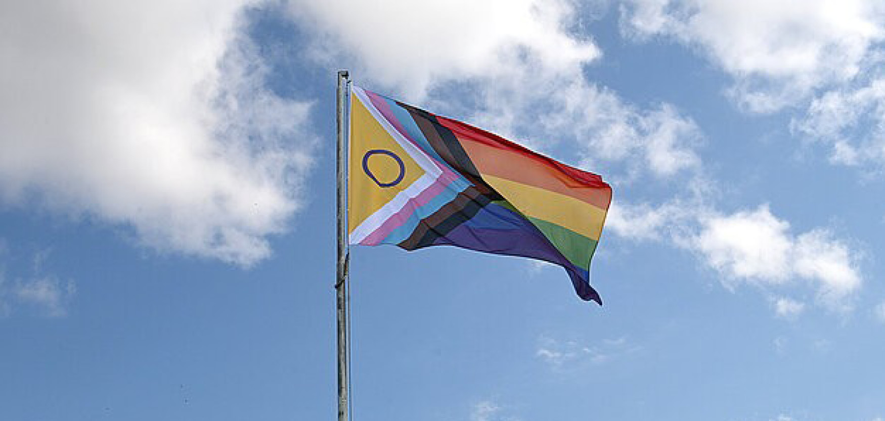 Regenbogenfahne mit zusätzlichen Farben weht an einem Fahnenmast vor blauem Himmel mit Wolken